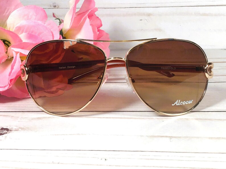 May include: Gold-framed aviator sunglasses with brown gradient lenses. The sunglasses have a double bridge and decorative accents on the temples. The text "Italian Design" and "Alcocer" are visible on the frame.