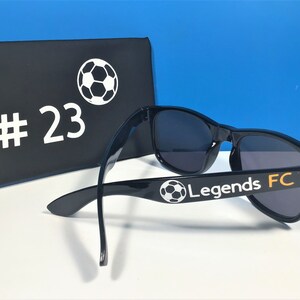 May include: Black sunglasses with a white soccer ball and the text "Legends FC" on the side. The sunglasses are in a black case with a white soccer ball and the number "#23" on the front.