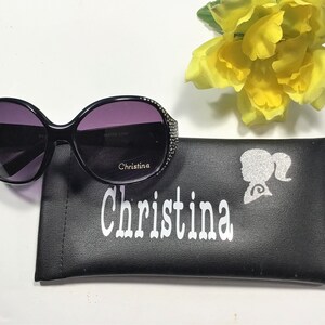 May include: Black sunglasses with a rhinestone trim and a black leather case with the name "Christina" printed on it in white.