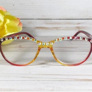 Puede incluir: Gafas con una montura degradada que pasa de un rojo intenso en los bordes exteriores a un amarillo transl&uacute;cido en el puente. El borde superior de la montura est&aacute; adornado con una fila de peque&ntilde;os diamantes de imitaci&oacute;n circulares.