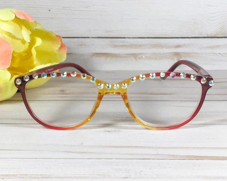Puede incluir: Gafas con una montura degradada que pasa de un rojo intenso en los bordes exteriores a un amarillo transl&uacute;cido en el puente. El borde superior de la montura est&aacute; adornado con una fila de peque&ntilde;os diamantes de imitaci&oacute;n circulares.