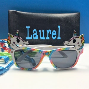 May include: White sunglasses with rainbow stripes and unicorn head details on the sides. The sunglasses come with a black case that says "Laurel" in blue letters.