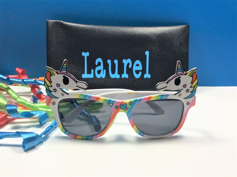 May include: White sunglasses with rainbow stripes and unicorn head details on the sides. The sunglasses come with a black case that says "Laurel" in blue letters.