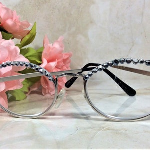 May include: Silver-framed eyeglasses with clear lenses, adorned with a row of sparkling rhinestones along the top rim. The glasses are set against a backdrop of pink flowers, creating a delicate and feminine aesthetic.