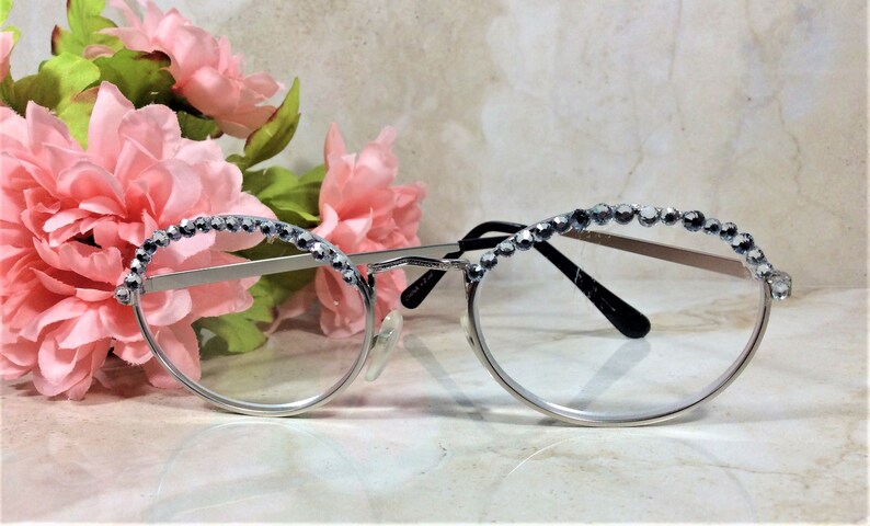 May include: Silver-framed eyeglasses with clear lenses, adorned with a row of sparkling rhinestones along the top rim. The glasses are set against a backdrop of pink flowers, creating a delicate and feminine aesthetic.