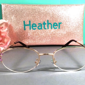 May include: Gold-rimmed spectacles with clear lenses and purple jewel accents. A rose gold glitter clutch with the name "Heather" in teal lettering is in the background. A pink flower is on the left side of the image.