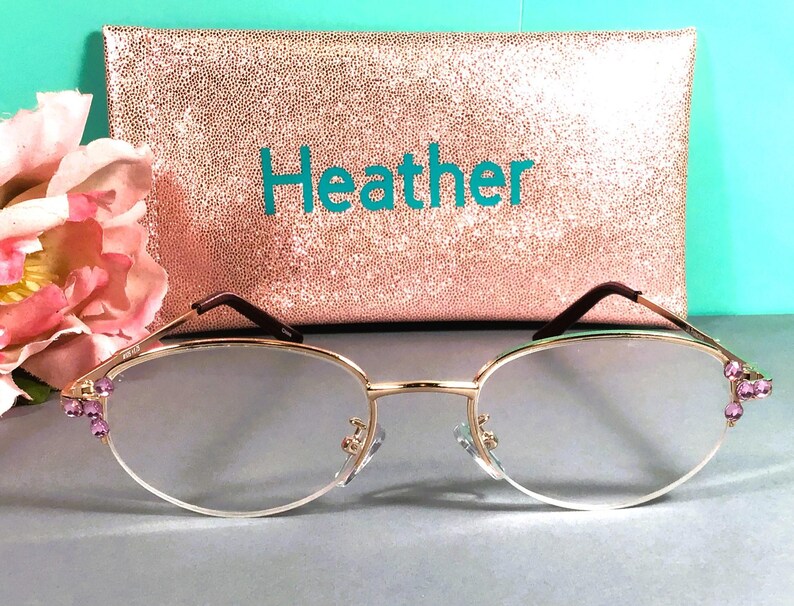 May include: Gold-rimmed spectacles with clear lenses and purple jewel accents. A rose gold glitter clutch with the name "Heather" in teal lettering is in the background. A pink flower is on the left side of the image.