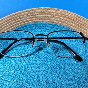 May include: Half-rimmed reading glasses with black frames, placed on a blue straw hat. The glasses feature a silver bridge and the text "R464 CHINA" and "+2.50". The hat has a woven pattern in brown and blue.
