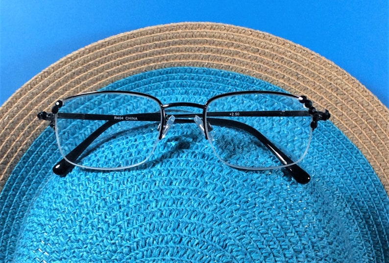 May include: Half-rimmed reading glasses with black frames, placed on a blue straw hat. The glasses feature a silver bridge and the text "R464 CHINA" and "+2.50". The hat has a woven pattern in brown and blue.