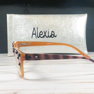 Op de afbeelding: Oranje montuurbril met decoratieve roze en zwarte versieringen op het montuur. De pootjes van de bril hebben een bruin schildpadpatroon. Een zilveren etui met de naam "Alexis" staat op de achtergrond.