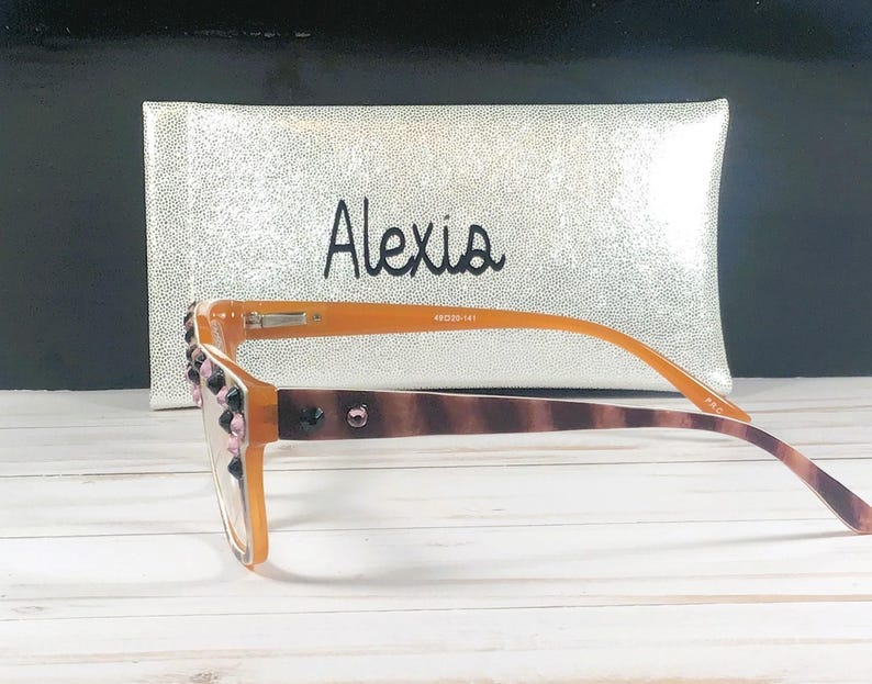 Op de afbeelding: Oranje montuurbril met decoratieve roze en zwarte versieringen op het montuur. De pootjes van de bril hebben een bruin schildpadpatroon. Een zilveren etui met de naam "Alexis" staat op de achtergrond.