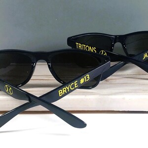 Puede incluir: Dos pares de gafas de sol negras con lentes oscuras. Las patillas presentan texto y gr&aacute;ficos en amarillo. Un par muestra TRITONS, un icono de b&eacute;isbol y BRYCE #13. El otro par muestra TRITONS, un icono de b&eacute;isbol y JAXSON #1 en letras amarillas.