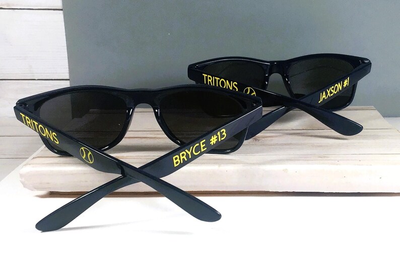 Puede incluir: Dos pares de gafas de sol negras con lentes oscuras. Las patillas presentan texto y gr&aacute;ficos en amarillo. Un par muestra TRITONS, un icono de b&eacute;isbol y BRYCE #13. El otro par muestra TRITONS, un icono de b&eacute;isbol y JAXSON #1 en letras amarillas.