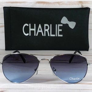 May include: Aviator sunglasses with silver frames and gradient blue lenses featuring the word Charlie printed on the right lens. The sunglasses are paired with a black rectangular case that displays the word Charlie and a bow icon in white.