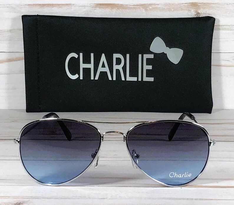 May include: Aviator sunglasses with silver frames and gradient blue lenses featuring the word Charlie printed on the right lens. The sunglasses are paired with a black rectangular case that displays the word Charlie and a bow icon in white.