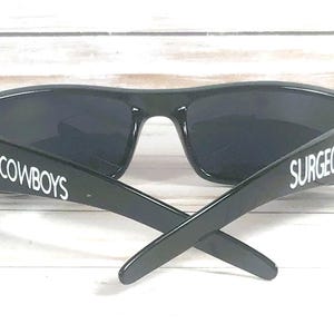 May include: Black sunglasses with dark lenses. The arms of the sunglasses have the word "COWBOYS" and "SURGEON" printed in white. A blue star is on the side of the frame.