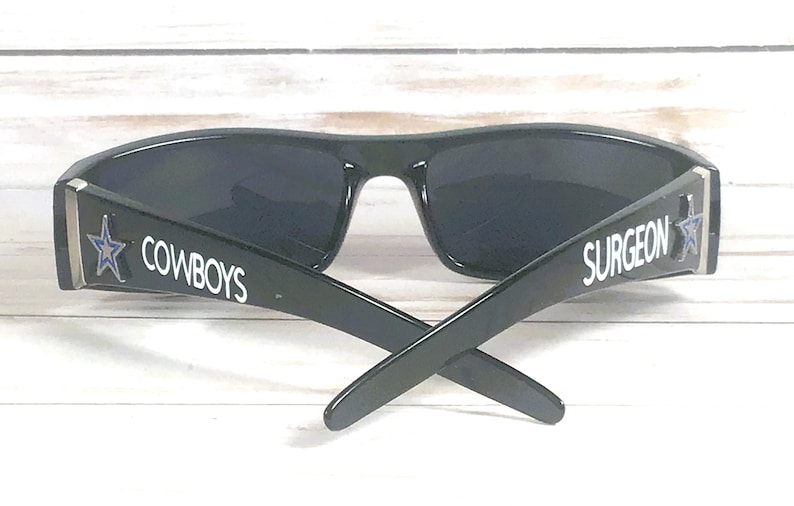 May include: Black sunglasses with dark lenses. The arms of the sunglasses have the word "COWBOYS" and "SURGEON" printed in white. A blue star is on the side of the frame.