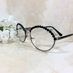 May include: Round-framed eyeglasses with a dark metal finish and black beaded accents along the top rims. The glasses are positioned near a cluster of white flowers, creating a stylish accessory.