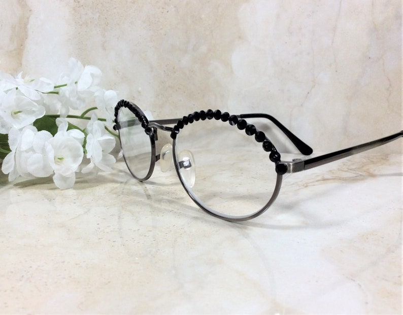 May include: Round-framed eyeglasses with a dark metal finish and black beaded accents along the top rims. The glasses are positioned near a cluster of white flowers, creating a stylish accessory.
