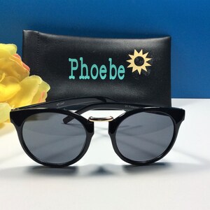 May include: Black round sunglasses with a gold accent on the bridge. The sunglasses are sitting on a white surface in front of a black case with the word "Phoebe" and a sun graphic.