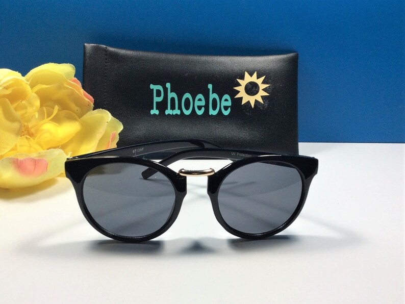 May include: Black round sunglasses with a gold accent on the bridge. The sunglasses are sitting on a white surface in front of a black case with the word "Phoebe" and a sun graphic.