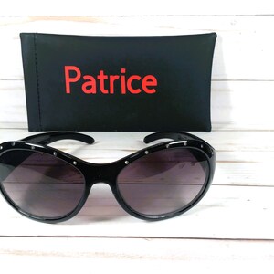 May include: Black sunglasses with dark tinted lenses and a frame featuring small embedded silver-tone studs along the top edge. Behind the sunglasses sits a black rectangular case with the word Patrice printed in red lettering on the front.
