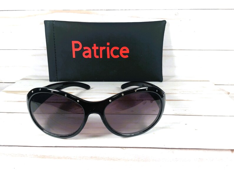 May include: Black sunglasses with dark tinted lenses and a frame featuring small embedded silver-tone studs along the top edge. Behind the sunglasses sits a black rectangular case with the word Patrice printed in red lettering on the front.