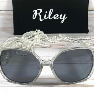 May include: Silver glitter sunglasses with a dark gray lens. The sunglasses are on a white surface with a string of white pearls.
