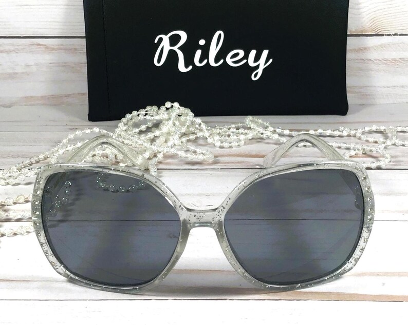 May include: Silver glitter sunglasses with a dark gray lens. The sunglasses are on a white surface with a string of white pearls.