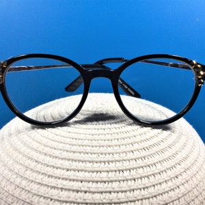May include: Black-framed eyeglasses with a round shape and gold accents on the temples. The glasses have clear lenses and are set against a blue background. The frame is made of a shiny black material.