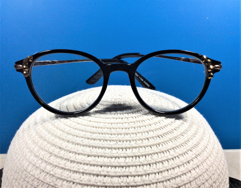 May include: Black-framed eyeglasses with a round shape and gold accents on the temples. The glasses have clear lenses and are set against a blue background. The frame is made of a shiny black material.