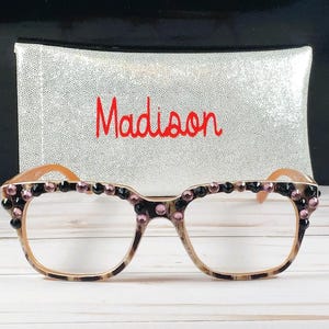 Op de afbeelding: Een bril met een luipaardprint montuur, versierd met zwarte en roze edelstenen. De bril staat voor een zilveren pailletten etui met de naam "Madison" in het rood geborduurd.