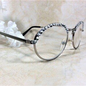 May include: Silver-framed eyeglasses with clear lenses. The upper rims are embellished with small, sparkling crystals. The temples are thin and dark, and the glasses are set against a soft, neutral background with white flowers.