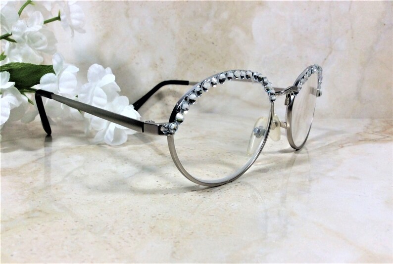 May include: Silver-framed eyeglasses with clear lenses. The upper rims are embellished with small, sparkling crystals. The temples are thin and dark, and the glasses are set against a soft, neutral background with white flowers.