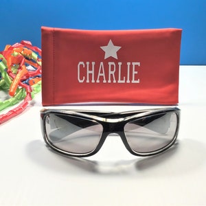 May include: A red sunglasses case with a white star and the name "CHARLIE" printed on it.  Black sunglasses with grey lenses are in front of the case.