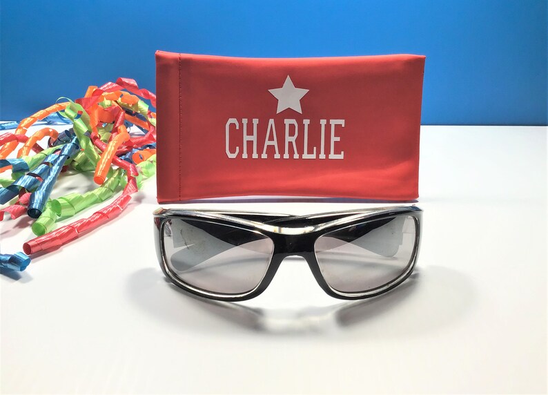 May include: A red sunglasses case with a white star and the name "CHARLIE" printed on it.  Black sunglasses with grey lenses are in front of the case.