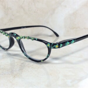 May include: Black-framed spectacles with a teal and yellow floral pattern. The top of the frames are adorned with small green gemstones. The glasses have a classic, elegant design.
