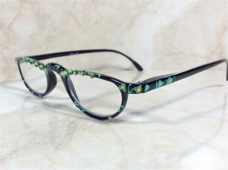 May include: Black-framed spectacles with a teal and yellow floral pattern. The top of the frames are adorned with small green gemstones. The glasses have a classic, elegant design.