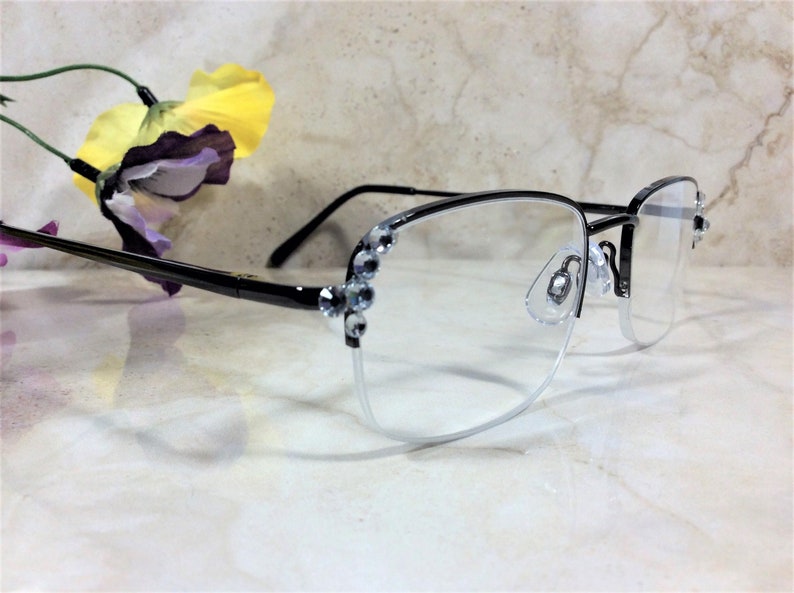 May include: Semi-rimless eyeglasses with a black frame and clear lenses. The upper frame is black, and the lower frame is a silver color. The temples are black, and the outer corners of the frame are decorated with clear rhinestones. A yellow and purple flower is in the background.