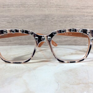 Puede incluir: Gafas de lectura con montura con estampado de leopardo, adornadas con brillantes transparentes en la parte superior. Las lentes son transparentes y las patillas son de color marr&oacute;n claro. El texto "RD-075 +1.50" est&aacute; impreso en la lente derecha.