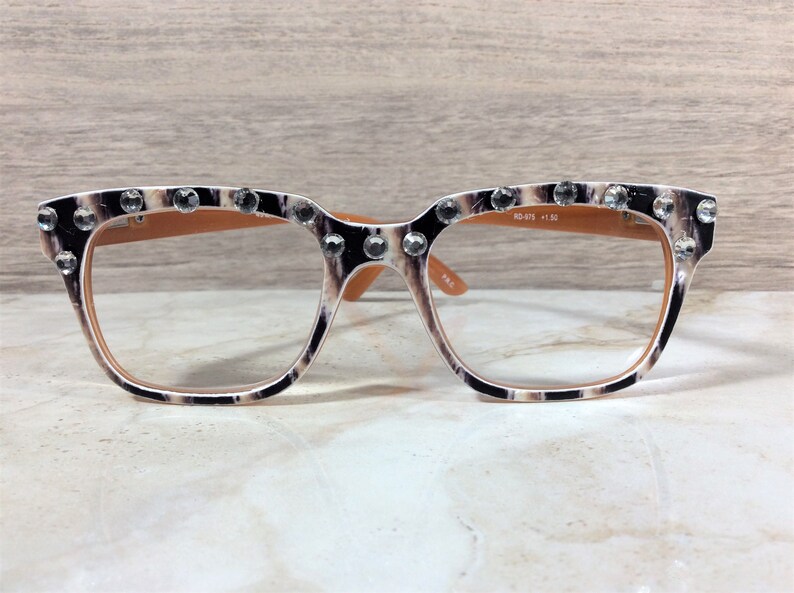 Puede incluir: Gafas de lectura con montura con estampado de leopardo, adornadas con brillantes transparentes en la parte superior. Las lentes son transparentes y las patillas son de color marr&oacute;n claro. El texto "RD-075 +1.50" est&aacute; impreso en la lente derecha.