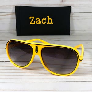 Puede incluir: Gafas de sol aviador amarillas con lentes oscuras. Detr&aacute;s de las gafas hay una funda negra con el nombre "Zach" bordado en amarillo. Las gafas tienen un ribete negro y un peque&ntilde;o emblema en el puente.