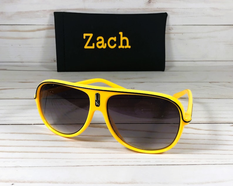 Puede incluir: Gafas de sol aviador amarillas con lentes oscuras. Detr&aacute;s de las gafas hay una funda negra con el nombre "Zach" bordado en amarillo. Las gafas tienen un ribete negro y un peque&ntilde;o emblema en el puente.