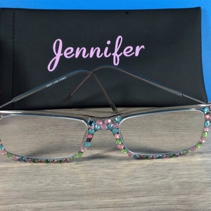 May include: Silver-framed reading glasses with colorful rhinestones along the top and bottom edges. A black case with the name "Jennifer" in pink script is behind the glasses. The glasses have a rectangular shape.