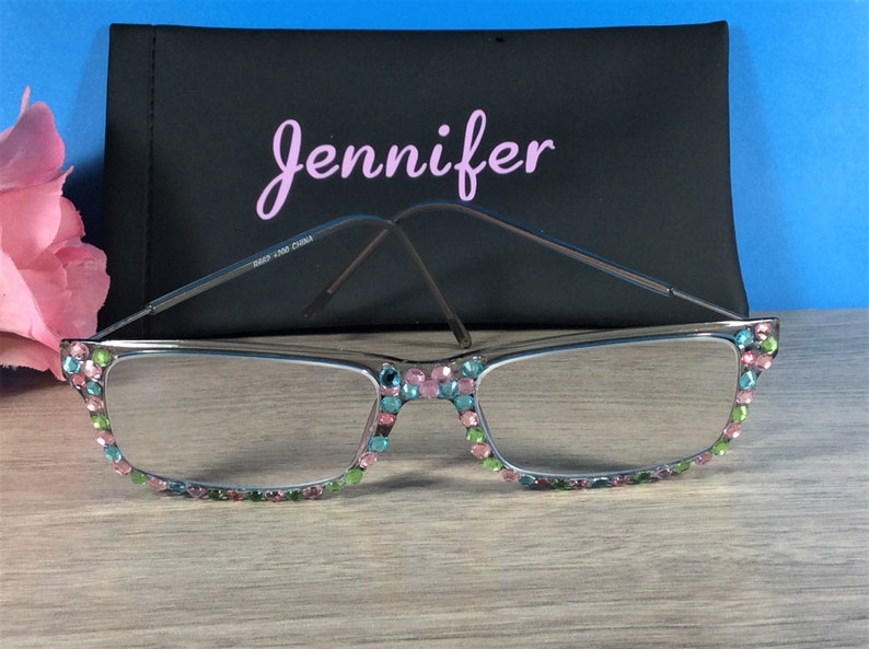 May include: Silver-framed reading glasses with colorful rhinestones along the top and bottom edges. A black case with the name "Jennifer" in pink script is behind the glasses. The glasses have a rectangular shape.