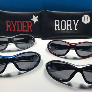 May include: Four pairs of sunglasses with black frames and gray lenses. The sunglasses are arranged in two rows of two. The top row has a pair of sunglasses with a black frame and a pair of sunglasses with a red frame. The bottom row has a pair of sunglasses with a blue frame and a pair of sunglasses with a black frame. The sunglasses are all in a protective case with the names "Ryder" and "Rory" printed on them.