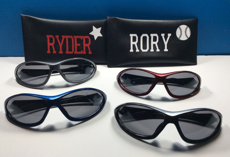 May include: Four pairs of sunglasses with black frames and gray lenses. The sunglasses are arranged in two rows of two. The top row has a pair of sunglasses with a black frame and a pair of sunglasses with a red frame. The bottom row has a pair of sunglasses with a blue frame and a pair of sunglasses with a black frame. The sunglasses are all in a protective case with the names "Ryder" and "Rory" printed on them.
