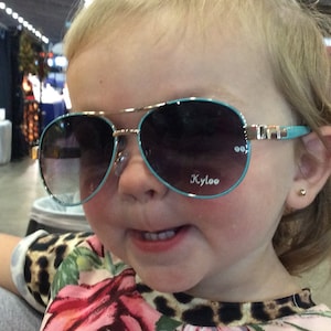 May include: A young child wearing blue and gold aviator sunglasses with the word "Kyloo" printed on the right lens. The child is wearing a leopard print and floral shirt.