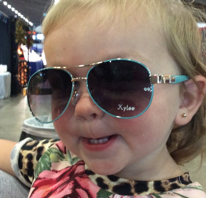 Pu&ograve; includere: Un bambino piccolo che indossa occhiali da sole aviator blu e oro con la parola "Kyloo" stampata sulla lente destra. Il bambino indossa una camicia con stampa leopardata e floreale.