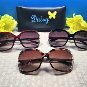 Op de afbeelding: Drie paar oversized zonnebrillen met verschillende kleuren frames. De frames zijn rood, zwart en schildpad. De zonnebrillen liggen op een wit oppervlak met een zwarte etui erachter. De etui heeft het woord "Daisy" en een vlinderontwerp erop.